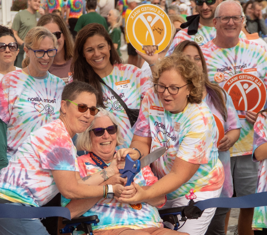 Person affected by ALS smiling while cutting a large ribbon with the help of others